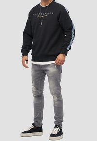 Sudadera negra con rayas azul claro en las mangas, con un logo amarillo. Combinada con jeans gris desgastados y zapatillas negras.