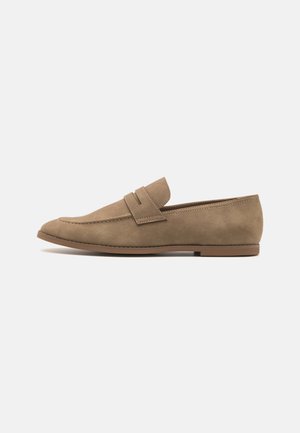 Lysebrun ruskind slip-on loafer med lav hæl og en syning detalje på tværs af vampen, vist mod en ensfarvet baggrund.