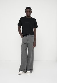 Camiseta negra con el texto "ORIGINAL" en relieve, combinada con jeans grises de pierna ancha con detalle de corbata y un ajuste relajado; zapatillas blancas.