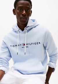 Hanorac albastru deschis cu șnururi, având inscripția "TOMMY HILFIGER EST. 985" în text închis la culoare pe piept. Materialul pare neted; croială lejeră.