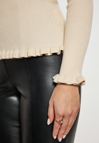 Cremfarbener gerippter Pullover mit gerafftem Saum und Ärmeln, kombiniert mit schwarzen Lederhosen. Die Hand ist mit einem goldenen Ring mit einem lila Stein verziert.