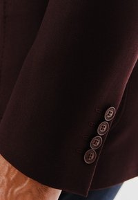 Rękaw burgundowej marynarki garniturowej z pięcioma brązowymi guzikami, gładką teksturą materiału, widocznym szwem i dopasowanym krojem.