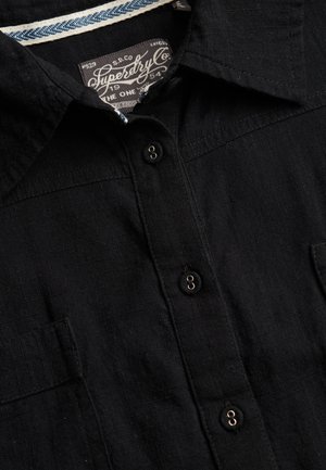 Close-up van een zwart overhemd met knopen, kraag en een zichtbaar Superdry-label aan de binnenkant van de halslijn.