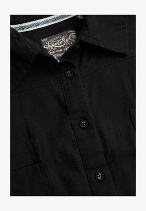 Close-up van een zwart overhemd met knopen, kraag en een zichtbaar Superdry-label aan de binnenkant van de halslijn.