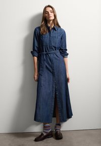 Vestido camisa de mezclilla en azul oscuro, con mangas largas, cierre de botones en la parte delantera, cinturón en la cintura y una abertura lateral. Combinado con mocasines y calcetines multicolores.