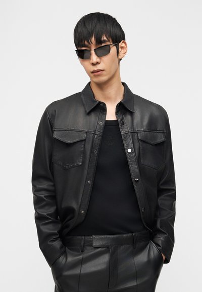 Άνδρας με μαύρο δερμάτινο μπουφάν, μαύρο πουκάμισο, μαύρο δερμάτινο παντελόνι και λεπτά μαύρα γυαλιά ηλίου, στέκεται με τα χέρια στις τσέπες.