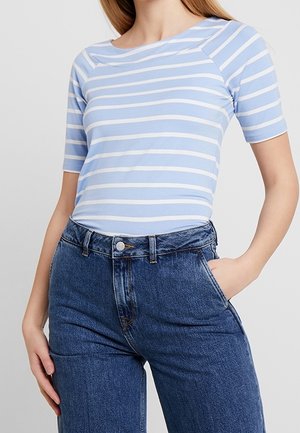 Mujer con una camiseta de manga corta a rayas azul claro y blanca metida en unos vaqueros azules de tiro alto, con una mano en el bolsillo.