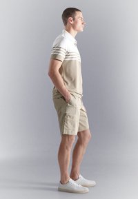 Hombre vestido con pantalones cortos cargo beige, camisa polo de rayas beige y blanca, zapatillas blancas, con las manos en los bolsillos, mirando hacia la derecha sobre un fondo liso.