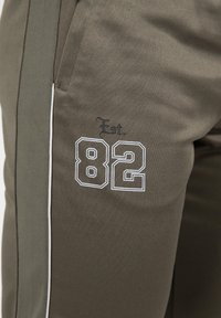 Olijfgroene sportbroek met witte accenten, met 'Est. 82' geborduurd aan de zijkant. Gemaakt van gladde stof, inclusief zakken.