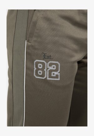 Olijfgroene sportbroek met witte accenten, met 'Est. 82' geborduurd aan de zijkant. Gemaakt van gladde stof, inclusief zakken.