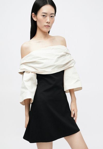 Crna haljina s uskim korzetom i lepršavom suknjom, koja uključuje off-white, naborani, oversized plašt preko ramena i rukave do tri četvrtine.