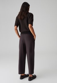 Maglione corto marrone a maniche corte abbinato a pantaloni scuri con una vestibilità rilassata. Le scarpe sono nere e hanno una finitura lucida, e ci sono tasche posteriori.