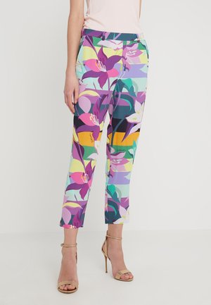 Kolorowe, kwiatowe spodnie o długości 7/8 noszone z beżowymi sandałami na wysokim obcasie i jasnym, bezrękawnym topem, stojące na jednolitym tle.