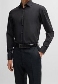 Camicia nera con bottoni e colletto classico, realizzata in tessuto liscio; abbinata a pantaloni scuri dalla trama ruvida e cintura nera.