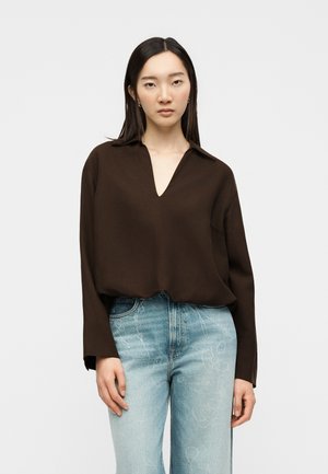 Femme aux longs cheveux foncés portant un t-shirt à manches longues marron foncé avec un col en V profond et un jean bleu clair taille haute avec un motif subtil.