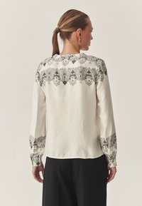 Blouse blanche en tissu lisse, ornée d'une broderie florale noire délicate sur les épaules et les poignets. Manches longues, coupe décontractée.
