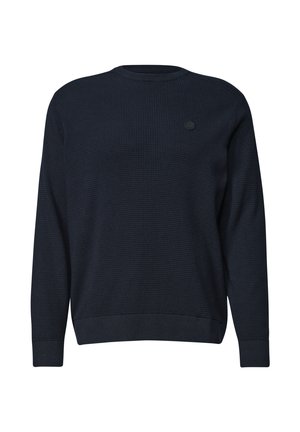 Mand med mørkt krøllet hår og skæg iført en marineblå sweater og lyseblå jeans, sidder afslappet op ad en hvid væg.
