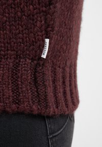 Burgunderfarbener Strickpullover mit dicker Textur und ribbeligem Saum; verfügt über ein kleines weißes Etikett mit dem Markennamen "MUSTANG."