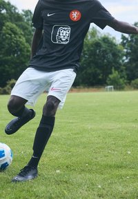 Svart Nike fotbollsjersey med prickmönster och orange lejonlogo, ihop med vita shorts med en rosa Nike-swoosh och svarta strumpor.