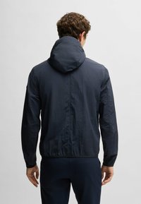 Marinblå lättviktsjacka med huva, med en krinklad yta och elastiska muddar. Designen har en figursydd silhuett och en justerbar nederkant.