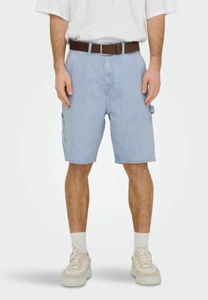 Mężczyzna w jasnoniebieskich dżinsowych szortach, białych tenisówkach, białych skarpetkach, brązowym pasku i białej koszulce na gładkim tle.