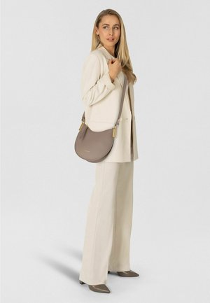 Vrouw in een crèmekleurig broekpak en taupekleurige hakken, die een taupekleurige crossbodytas met goudkleurige details vasthoudt en opzij kijkt tegen een effen achtergrond.