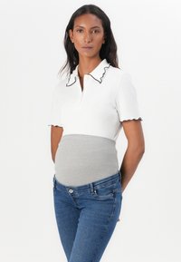 White polo shirt with ruffled black collar and cuffs, paired with grey stretchy waistband. Model wearing blue fitted jeans stands with hands on hips.