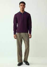 Bordó cipzáras pulóver tengerészkék gallérral, barna kockás nadrággal és fekete cipővel, semleges háttér előtt állva.