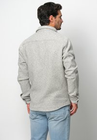 Grau strukturierte Jacke mit Kragen, sichtbaren Nähten und geknöpften Manschetten, kombiniert mit hellblauen Jeans, von hinten betrachtet.