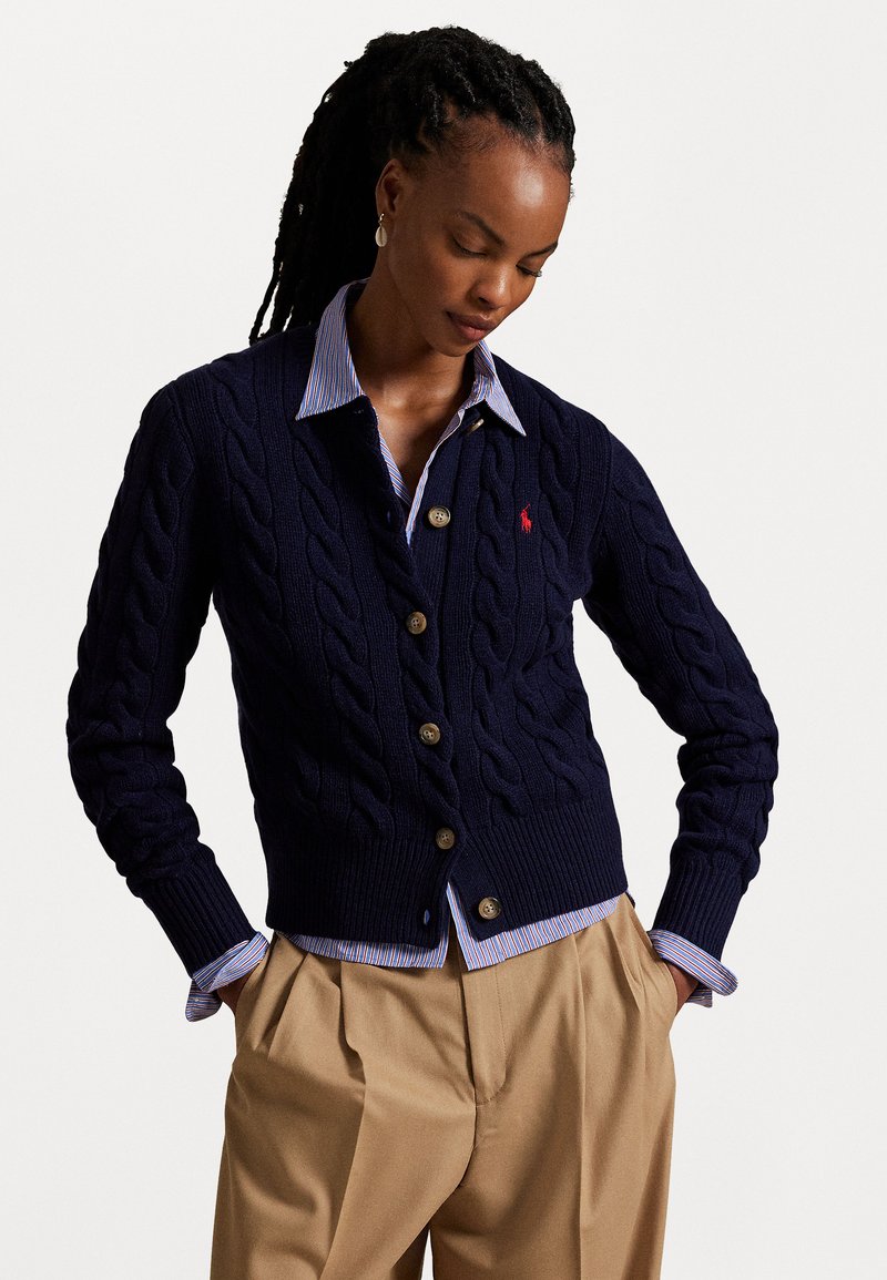 Woman with braided hair wearing a navy cable-knit cardigan over a striped shirt and beige pleated pants, hands in pockets, looking down.