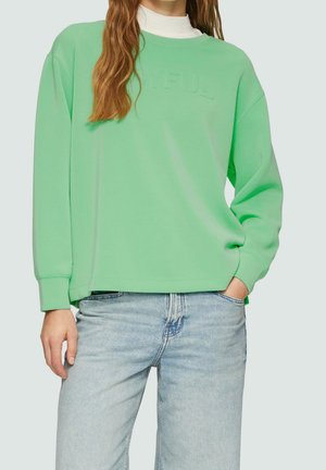 Personne portant un sweat-shirt oversize vert clair avec un texte embossé discret et un jean bleu clair, une main dans la poche et de longs cheveux visibles.
