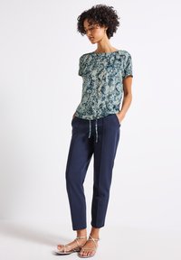 Helles, blaues, schlangenbedrucktes T-Shirt mit kurzen Ärmeln, kombiniert mit navyblauen, schmal geschnittenen Hosen mit elastischem Kordelzugbund und seitlichen Taschen. Silberne, Riemchensandalen.