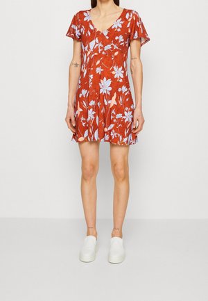 Vrouw draagt een rode bloemenjurk met korte mouwen met blauwe bloemen en witte instapschoenen, staand tegen een effen witte achtergrond.