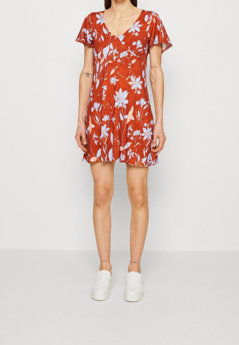 Femme portant une robe rouge à fleurs à manches courtes avec des fleurs bleues et des chaussures blanches à enfiler, debout devant un fond blanc uni.