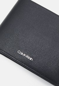 Must värvitud rahakott, mis on valmistatud sünteetilisest materjalist, millel on hõbedane "Calvin Klein" logo ja kihiline disain. Omab äärte ümber õmblusi.