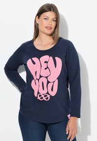 Camisa de manga larga navy con un gráfico texturizado en rosa que dice "HEY YOU" en el frente. Cuello redondeado y ligera abertura lateral. Llevada con vaqueros azules.