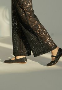 Pantalones de encaje negro con perneras anchas, que presentan intrincados patrones florales, combinados con bailarinas negras con purpurina, con punta redonda y tira al tobillo.
