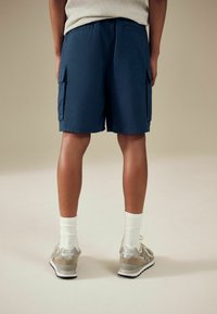 Pantaloni cargo blu con due tasche laterali, abbinati a sneakers beige. Calzini bianchi a coste sono visibili sopra la linea delle scarpe.