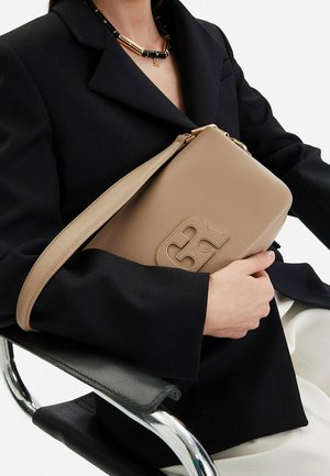 Osoba v černém saku sedící na chromové židli drží béžovou koženou kabelku se zlatým logem a páskem přes paži, nosí černo-zlatý náhrdelník.