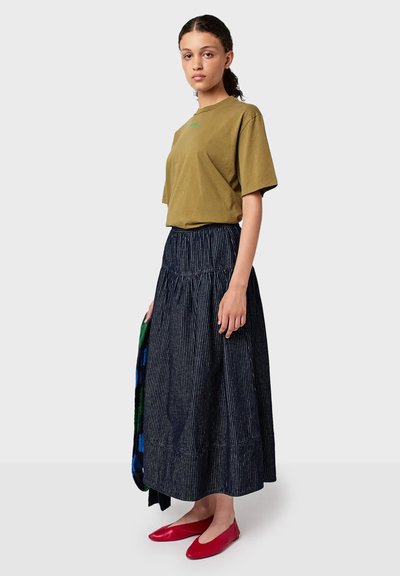 Donna in piedi che indossa una maglietta verde oliva, una lunga gonna a righe scure, scarpe rosse, e tiene in mano un oggetto piegato a scacchi multicolori.