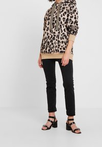 Hoodie beige con stampa leopardata e dettagli dorati, tasca frontale, abbinato a pantaloni neri e tacchi neri con lacci. Tessuto morbido e vestibilità rilassata.