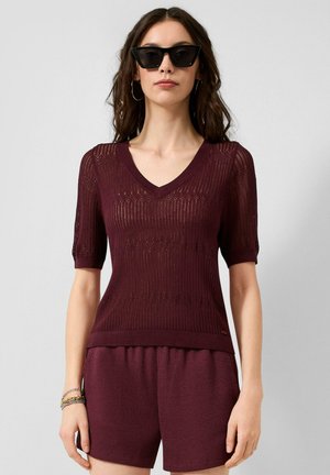 Frau mit Sonnenbrille und Creolen-Ohrringen, einem kurzärmeligen, strukturierten bordeauxroten Strickoberteil und passenden Shorts vor einem einfarbigen Hintergrund.