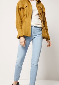 Senapsgul, texturerad jacka med stora fickor och knappar, i kombination med ljusblå slim-fit jeans. Svarta slip-on skor fulländar looken.