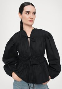Svart bluse med lange pufferm, samlet tekstur og innsnevret midje. Har et båndfeste i nakken og en avslappet passform.