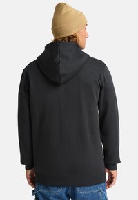 Schwarzer Kapuzenpullover mit lockerer Passform, weichem Stoff und gerippten Bündchen. Getragen mit einer hellbraunen Mütze. Einfaches, minimalistisches Design.