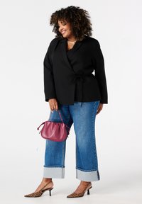 Zwarte wikkeljas met een strik in de taille, gecombineerd met wijde denim jeans met omgeslagen pijpen. Geaccessoireerd met een kleine roze tas en luipaardprint hakken.