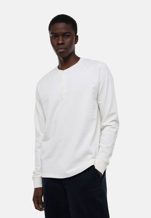 Jeune homme portant un henley blanc à manches longues avec des boutons, debout, une main dans la poche d'un pantalon noir, fond neutre.