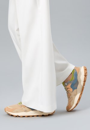 Ben iført brede hvide bukser og flerfarvede beige, blå og grønne sneakers med teksturerede brune såler mod en grå baggrund.