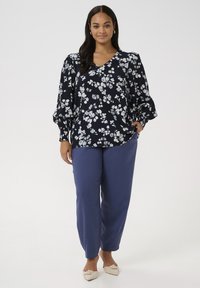 Bluză florală bleumarin cu mâneci bufante, decolteu în V; asociată cu pantaloni albaștri deschis, croiți, și pantofi plată crem cu un nod decorativ.