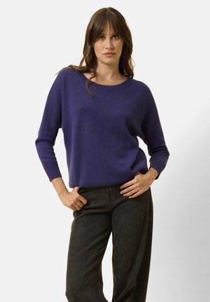 Maglione viola con scollatura rotonda, maniche lunghe e vestibilità rilassata, abbinato a jeans neri a gamba larga. Texture liscia, accenti minimi.
